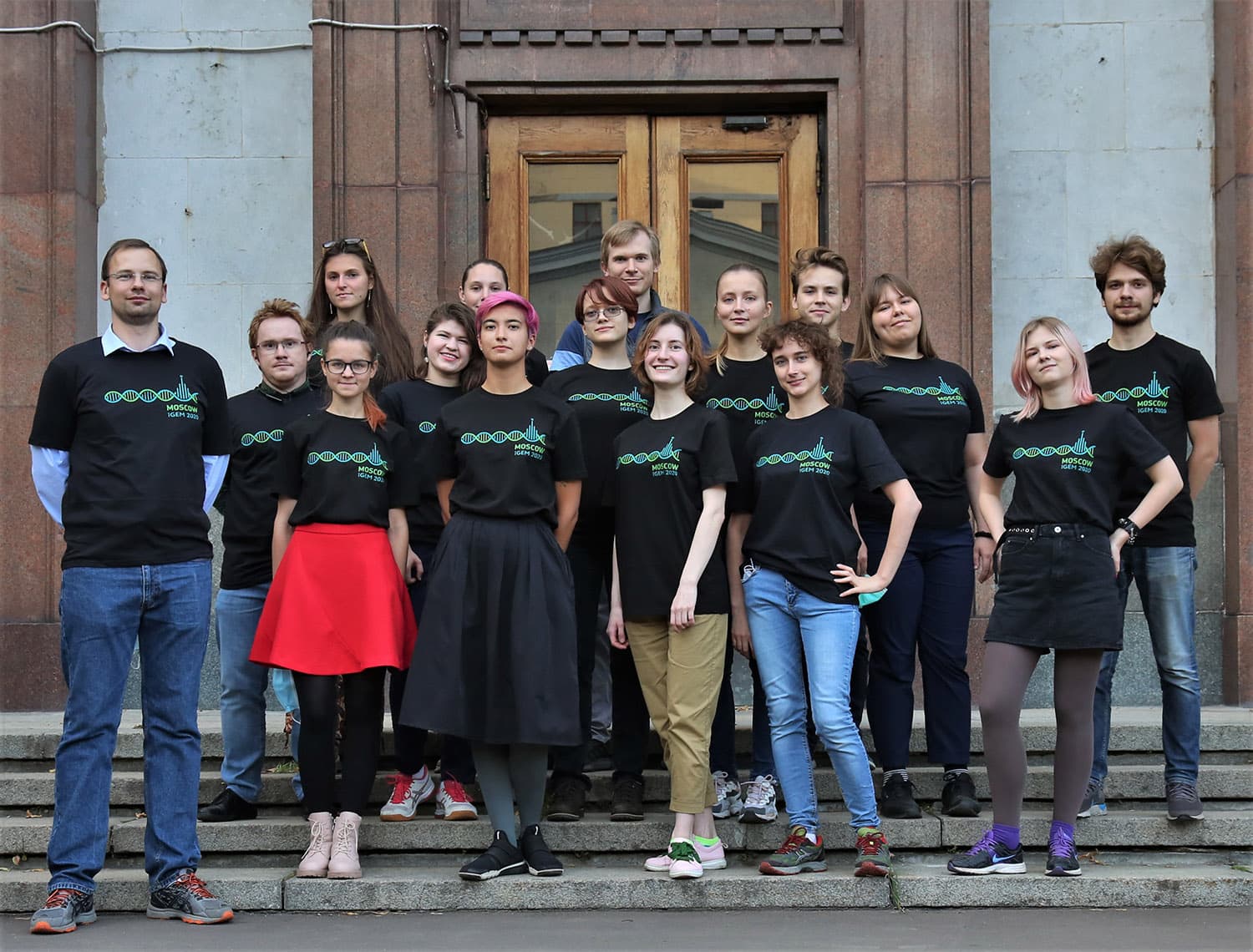 Group photo of the iGEM 2020 gold-medalist team Moscow, all wearing matching black t-shirts printed with a DNA helix graphic and "MOSCOW iGEM 2020", standing on the steps outside their university building